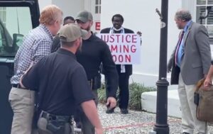 PHOTO Preacher Standing Outside Courthouse Door With Sign That Says Justice Coming Soon While Alex Murdaugh Is Escorted From Police Car Inside